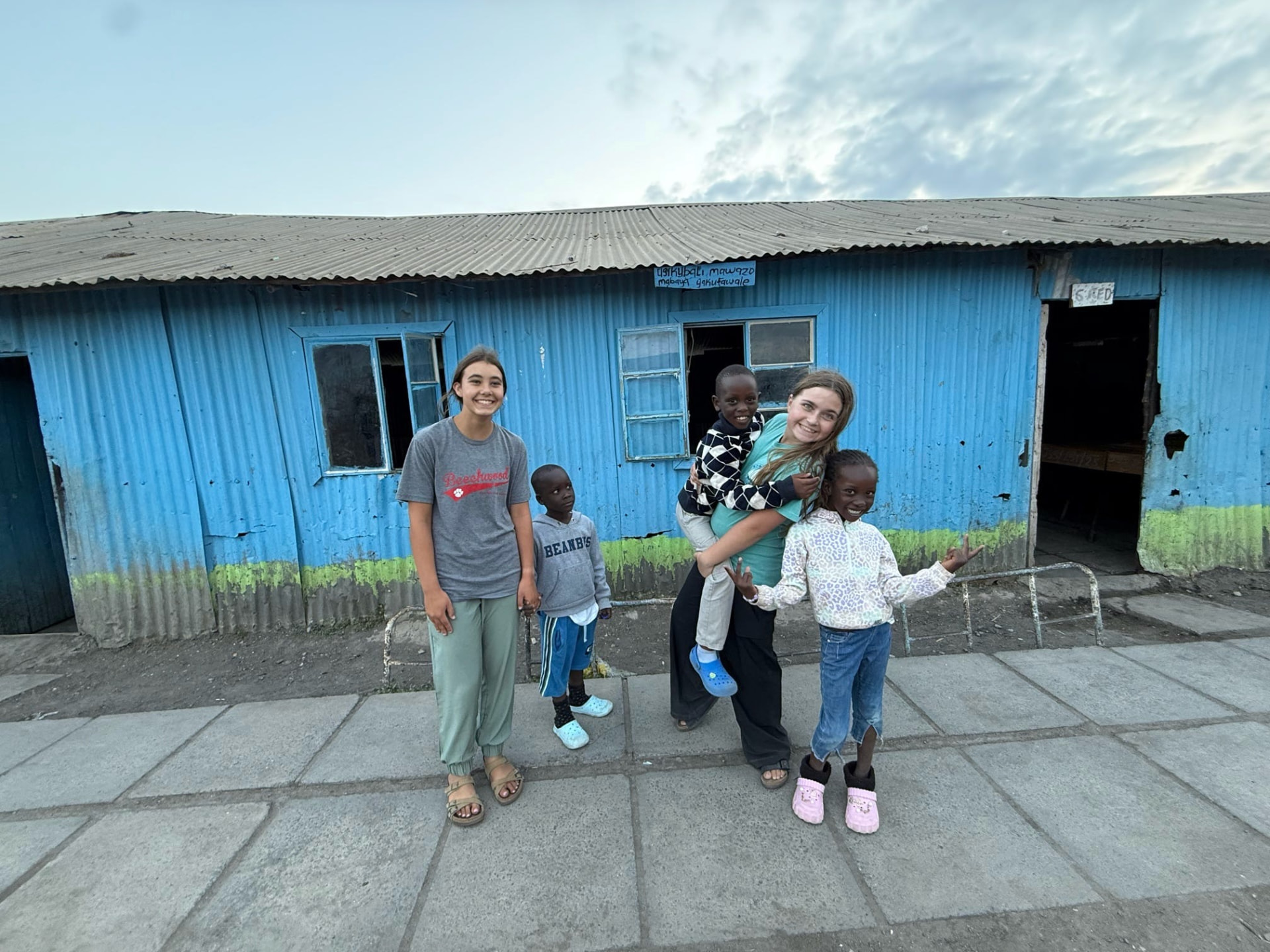 Two team members standing with three young children outside a blue corrugated iron classroom