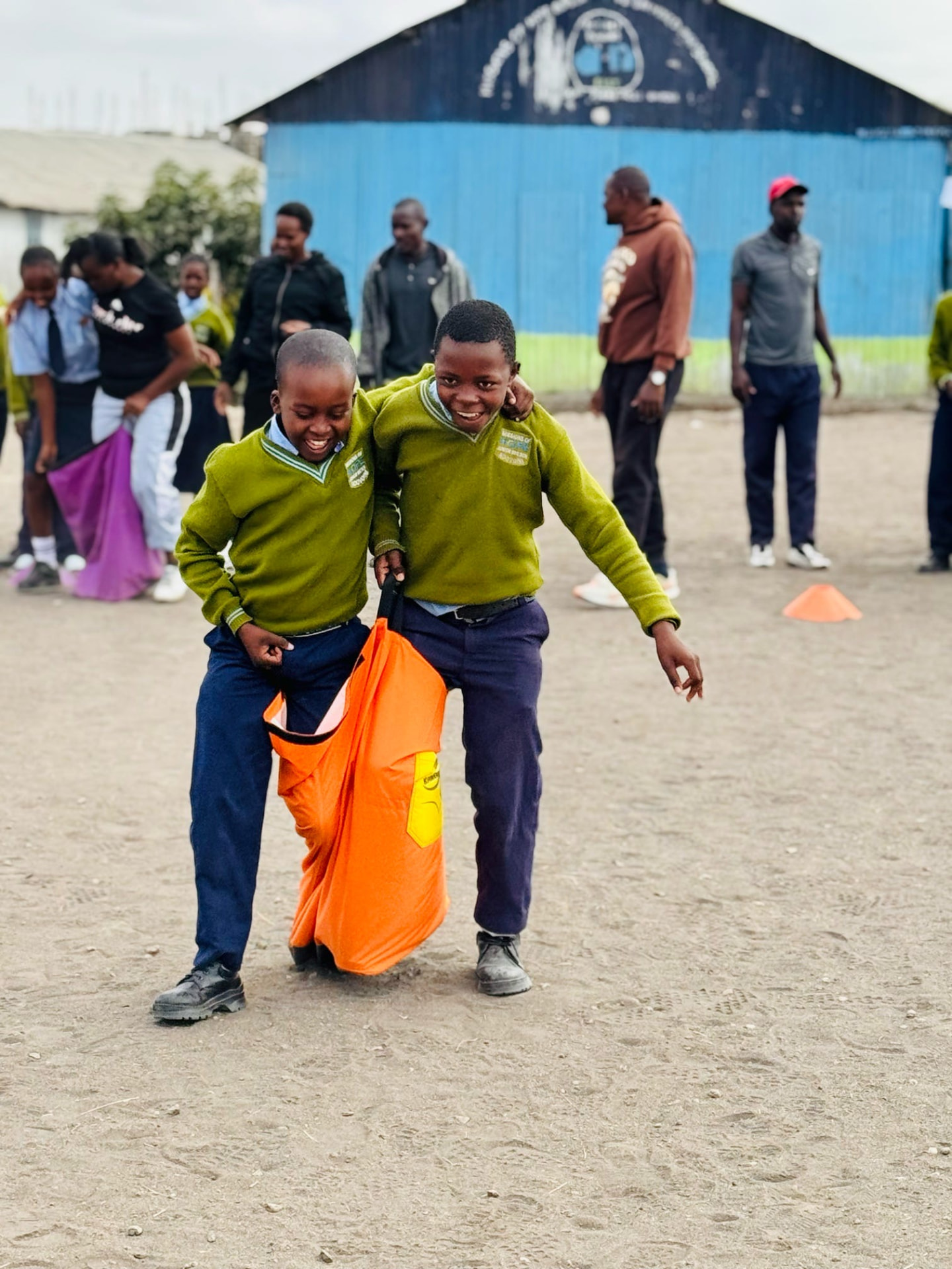 Two students in green sweaters laughing through a sack race on the school grounds
