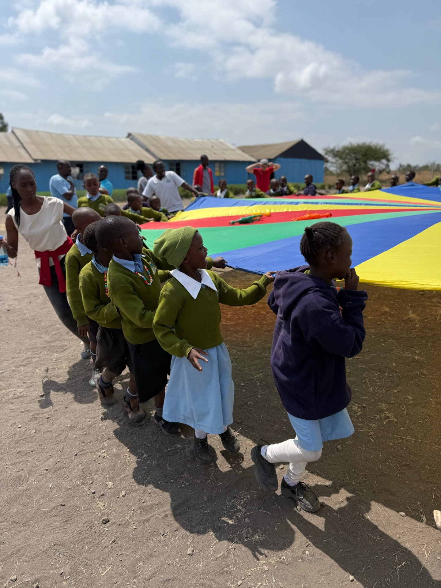 Young students in green sweaters pulling a colorful parachute across a dirt field